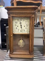 Front view of wooden wall clock with glass door closed, showing cream clock face with Arabic numerals and brass pendulum visible through glass.
