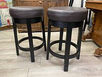 Two modern swivel bar stools with dark brown leather round padded seats and black wooden bases, shown from the side and front angles.