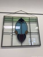 Front view of the rectangular stained glass window panel showing multicolored glass pieces arranged in a grid with a floral or abstract central design and metal frame with hanging chain.