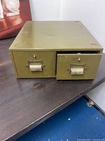 Metal card catalog filing system with two drawers on a table, showing wear and olive green color.