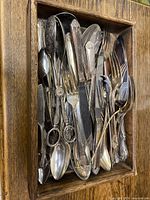 Overhead photo of mixed silverware pieces inside a wooden box showing silver-plated ornate forks, knives, and spoons stacked together with visible tarnish.