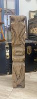 Full view of primitive tiki wood carving showing stylized facial and body features, natural wood finish, standing on floor against trunk and furniture in background.