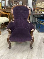 Front view of a purple velvet upholstered antique armchair with tufted backrest, wooden frame and gold trim accents.