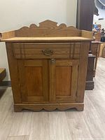 Front view of rustic wooden cabinet showing distinctive carved top and scalloped bottom edge, single drawer with metal handle, and two closed doors.