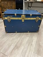Front view of the blue storage trunk showing central brass lock, corner reinforcements, and side brass accents