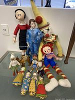 Overall view of assorted dolls and figurines including large crochet clown, rag dolls, wooden and ceramic figures, and cultural dolls.