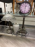 Photo showing vintage desk lamp, decorative vase, metal clock with Roman numerals, and metal wall mounted candlestick holders all arranged on a metal shelf.