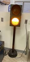 Front view of vintage traffic light in a room, lower amber light illuminated, showing metal pole base and electric cord.