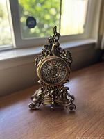 Full front view of brass antique mantle clock showing floral and paisley detailing with Roman numerals on dial.