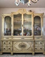 Full frontal view of the cream-finished cabinet showing upper glass doors, lower storage doors, carved crest and bun feet.