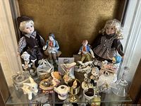 Wide view of porcelain dolls and assorted figurines displayed on a glass shelf, featuring the two dolls and multiple small porcelain pieces.