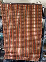 Full view of one woven wood shade hanging, showing multicolor patterned woven wood strips in vertical stripes including orange, brown, and beige tones.
