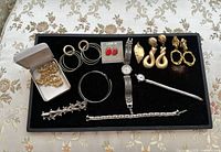 Full display of jewelry lot with multiple earrings, bracelets, and watches on black tray over floral fabric.