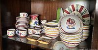 Shows overall collection of ceramic dishware including small stacked plates, mugs, bowls, a pitcher, and serving dishes with rooster and floral hand-painted designs.