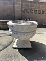 Side view of the large vintage concrete Roman-style planter on a paved outdoor surface showing the detailed decorative relief around the rim and pedestal base.