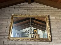 Large rectangular mirror mounted on a brick wall, reflecting a chandelier and ceiling beams.