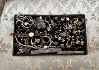 Top view of a black tray filled with a variety of vintage jewelry and coins on floral fabric background.