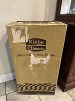 Closed cardboard box labeled Kirby Classic with signs of wear and tape residue.
