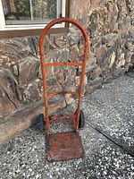 Orange painted metal hand truck dolly showing front with rectangular base plate, tubular handle, and two black wheels, placed on stone patio.