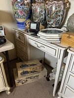 Photo shows the vintage white French Provincial desk with three drawers. On top and around the desk are various items such as large floral vases, a small TV monitor, framed glass pieces, and vintage toy boxes.