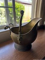 Front angled view of vintage brass coal scuttle ash bucket showing curved spout, round base, and metal handle with porcelain grips near a window.