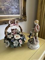 Photo showing both Capodimonte porcelain items: the floral bouquet basket and the boy figurine with dog on a surface.