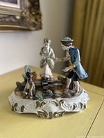 Overall side view of the ceramic sculpture showing woman, man, and dog with ornate base and gold detailing.