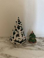Photo showing both ceramic Christmas tree figures on marble surface against plain wall