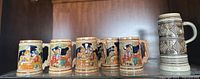 Front view of six vintage ceramic beer steins showing colorful painted German figures and one textured carved stein