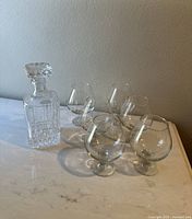 Full set showing one crystal decanter with stopper and six brandy snifter glasses on a marble surface against a plain wall.