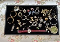Top view of a black felt tray containing assorted vintage jewelry including earrings in various styles, pins, rings, and one red strap watch.
