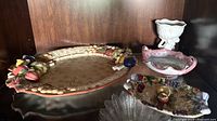 Photo showing large oval ceramic tray with fruit decoration, small pink scalloped bowl, small colorful fruit ceramic dish, and white pedestal dish on a wooden shelf