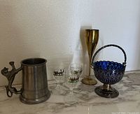 Group shot of all items on marble surface against white wall. Items shown from left to right: metal beer mug with cat handle, two small decorated glass cups, tall gold goblet, silver-tone basket with blue glass.