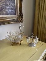 Glass swan dish and hanging crystal grape salt and pepper holder set on yellow surface with framed wall art background.