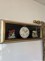 Framed shadow box display mounted on wall showing two porcelain bird figurines and a clock face between them.