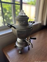 Full view of vintage metal coffee maker with glass lid, wooden handles, and spout on wooden table near window.