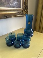 Blue hand blown glass pitcher and five matching tumblers on yellow table in indoor setting.