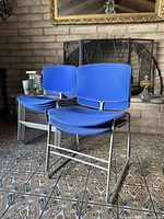 Two blue Steelcase stacking chairs shown side by side on patterned tile floor with brick fireplace background.
