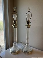 Pair of vintage mid-century table lamps on a white surface near a window, showing full lamp height and clear glass and brass-tone metal detail.