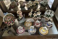 Full view of all items including demitasse cup and saucer sets, floral planters, and porcelain figurines on mirrored surface.