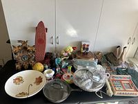 Full overview of the lot showing skateboard, large ceramic bowl, clock, toys, slippers, and other items arranged on a table