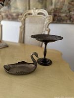 Vintage swan-shaped trinket dish and ornate pedestal dish shown together on a table with vintage chairs in the background.