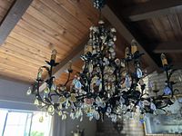 Full view of two-tier chandelier from below against wooden ceiling and window, showing metal frame, porcelain flowers, and hanging iridescent glass drops.