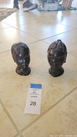 Front view of two dark brown ebony wood carved head statues, a man and a woman, on tiled floor with a lot number tag visible.