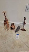 Four handcrafted African themed decorative art pieces displayed on floor: wooden giraffe sculpture, painted vase, stone mask sculpture, and wooden figurine holding spear with beads.