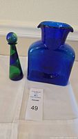 Full view of the cobalt blue glass water bottle and the smaller blue-green decorative decanter with stopper on a white cloth.