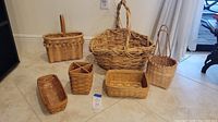 Group photo of six woven baskets in various shapes and sizes arranged on tile floor near wall.
