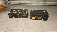 Pair of two black vintage storage trunks placed on a concrete floor, showing wear and labels.