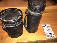 Two cylindrical black leather carrying cases for camera lenses, one marked with seller's tag noting Hanimex 300mm and Petri 135, showing signs of wear.