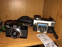 Photo showing Kodak Instamatic X-25 camera next to Petri Color 35 camera and black carrying case on wooden shelf